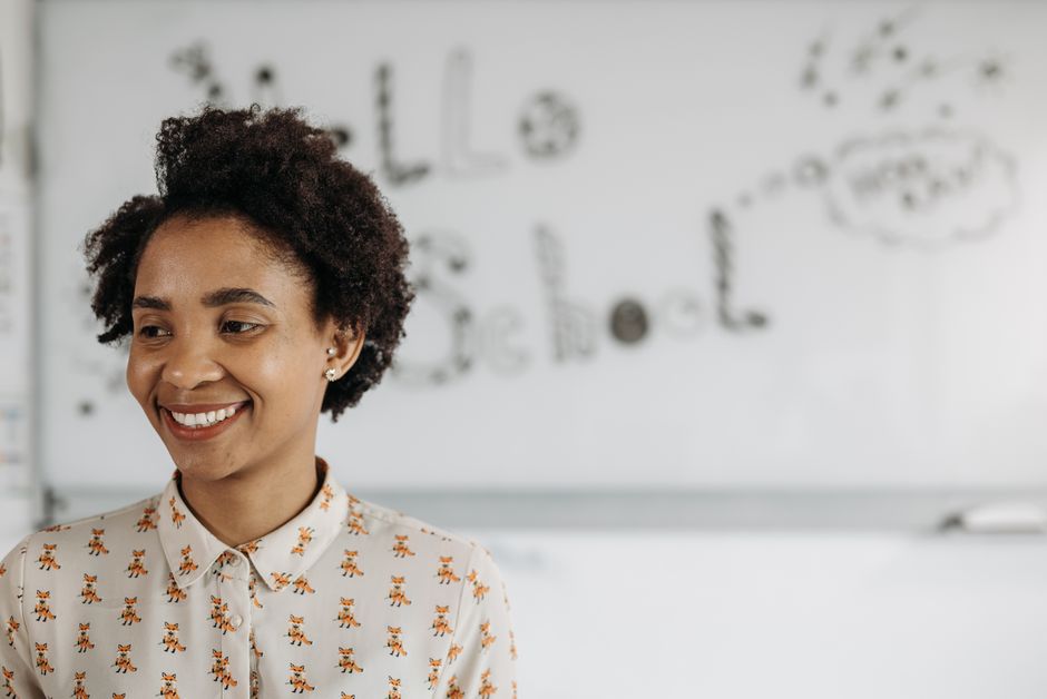 A friendly female teacher smiles in front of a classroom whiteboard.