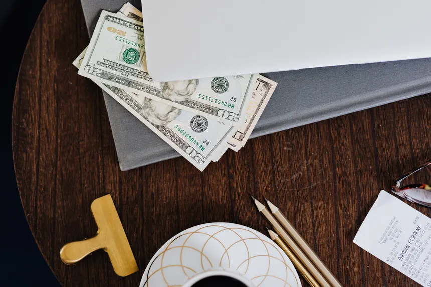 Stylish workspace featuring coffee cup, dollar bills, laptop, and stationery on a wooden table.