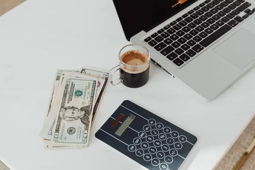 A modern workspace featuring a calculator, cash, and laptop with coffee.