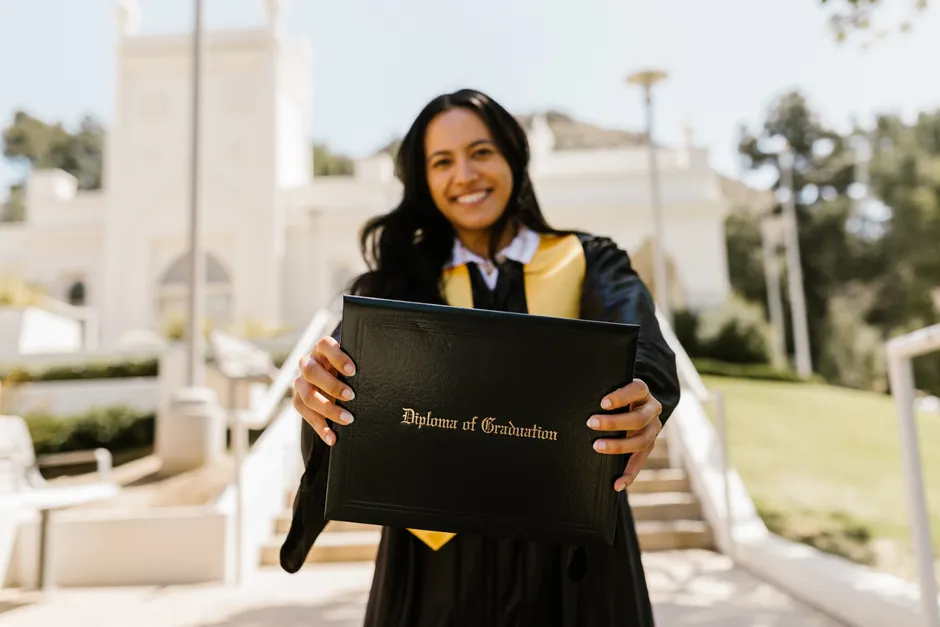 Smiling graduate holding a diploma outdoors in a sunny setting.