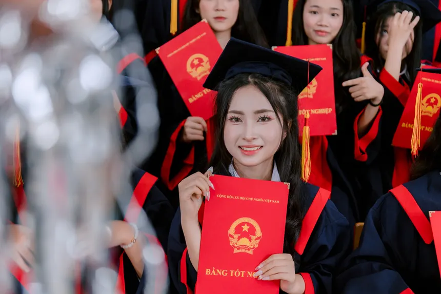 A cheerful female graduate holds a diploma during a graduation ceremony, surrounded by fellow graduates in caps and gowns.
