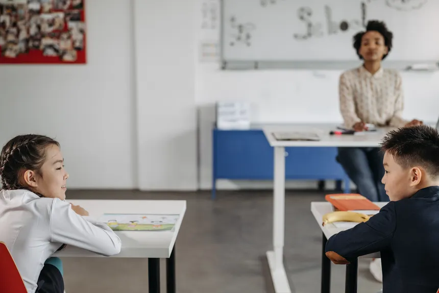 Two students engage in a classroom setting with their teacher in the background.