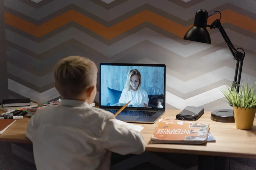 A child participates in an online lesson from home, using a laptop for video conferencing with a teacher.