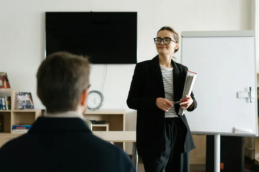 Caucasian female teacher in black blazer holding books, presenting to a student in a classroom.