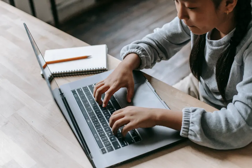From above of crop concentrated Asian schoolgirl wearing domestic clothes sitting at table with copybook while typing on wireless laptop in light room