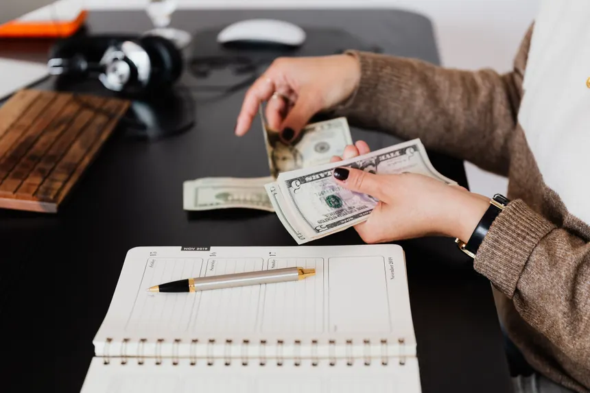 Side view of crop anonymous smartly dressed female entrepreneur counting dollar banknotes while sitting at modern office desk with planner and pen