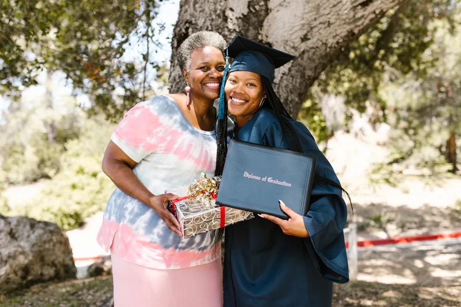 Graduating student with family member celebrating under a tree with a diploma and gift.